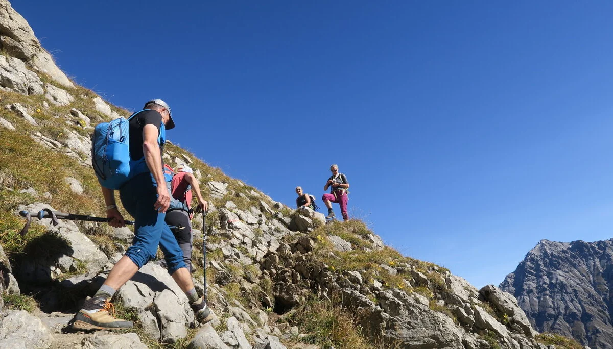 Wanderer mit Rucksack und Kappe steigen auf felsigem, grasbewachsenem Berghang unter blauem Himmel auf. | © DAV-FN / I. Eichenberg