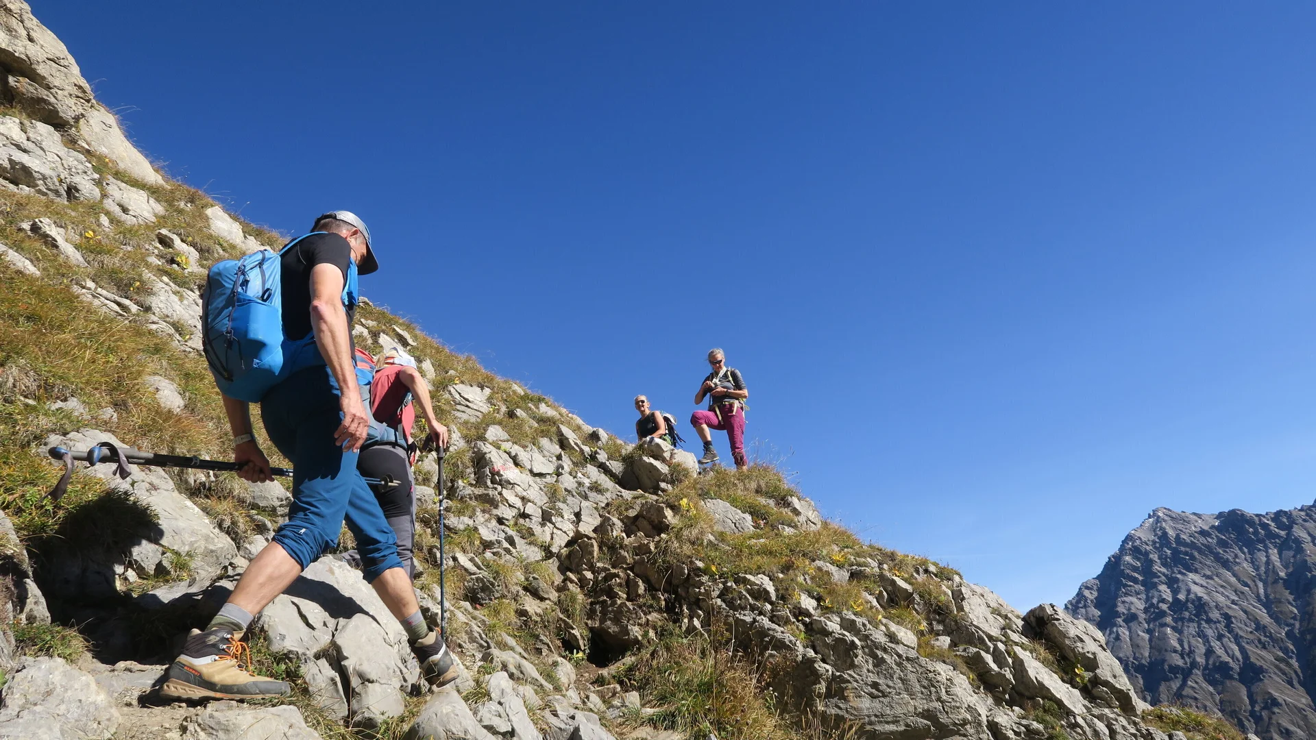 Wanderer mit Rucksack und Kappe steigen auf felsigem, grasbewachsenem Berghang unter blauem Himmel auf. | © DAV-FN / I. Eichenberg
