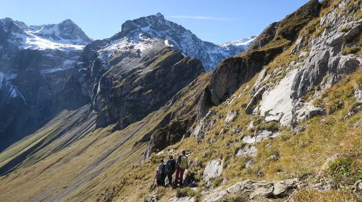 Berglandschaft mit schneebedeckten Gipfeln, steilem Gras- und Felsabhang und drei Wanderern auf einem Pfad. | © DAV-FN / I. Eichenberg