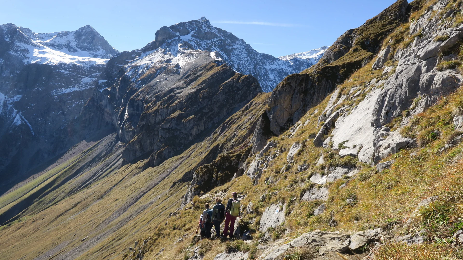 Berglandschaft mit schneebedeckten Gipfeln, steilem Gras- und Felsabhang und drei Wanderern auf einem Pfad. | © DAV-FN / I. Eichenberg