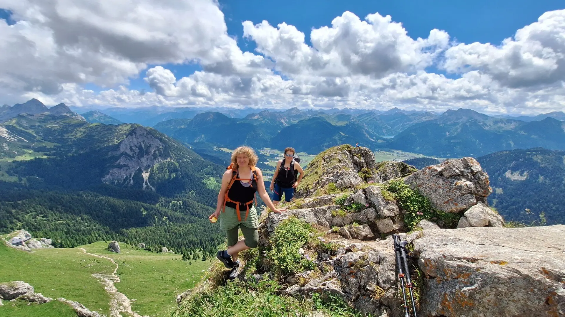 Zwei Wandernde steigen auf felsigem Pfad in den Bergen, umgeben von grünen Wiesen und leicht bewölktem Himmel | © DAV-FN / Karin B., Silke S., Robert H.
