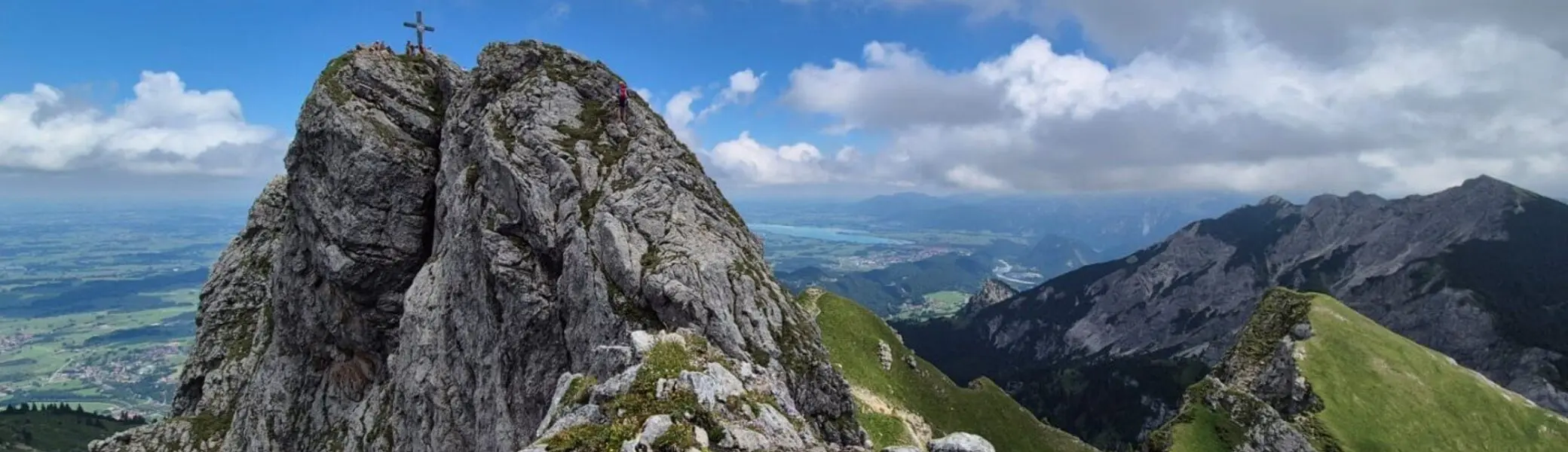 Felsgipfel mit Gipfelkreuz am Aggenstein, umgeben von grünen Bergwiesen und bewölktem Himmel | © DAV-FN / Karin B., Silke S., Robert H.