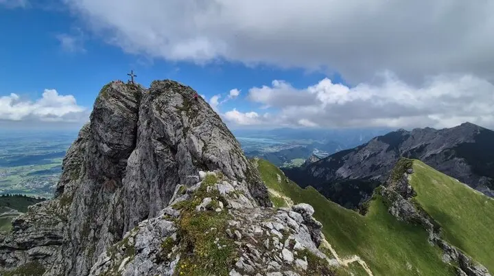Felsgipfel mit Gipfelkreuz am Aggenstein, umgeben von grünen Bergwiesen und bewölktem Himmel | © DAV-FN / Karin B., Silke S., Robert H.