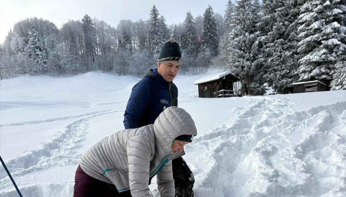 Zwei Personen knien im Schnee vor einem verschneiten Wald und einer Holzhütte, eine Person übt die Feinsuche mit dem LVS-Gerät | © DAV-FN / N. Gersten