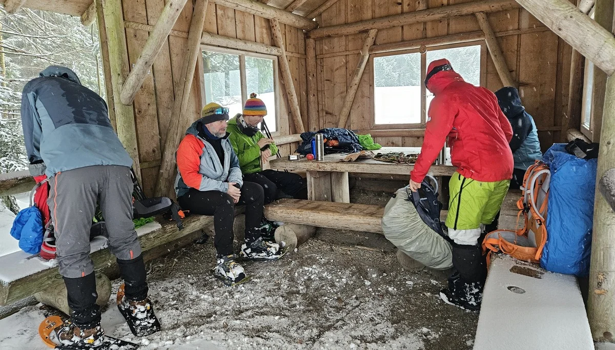 Mehrere Personen in Winterkleidung mit Schneeschuhen sitzen und stehen in einer hölzernen Schutzhütte mit Schneeboden | © DAV-FN / A. Schababerle