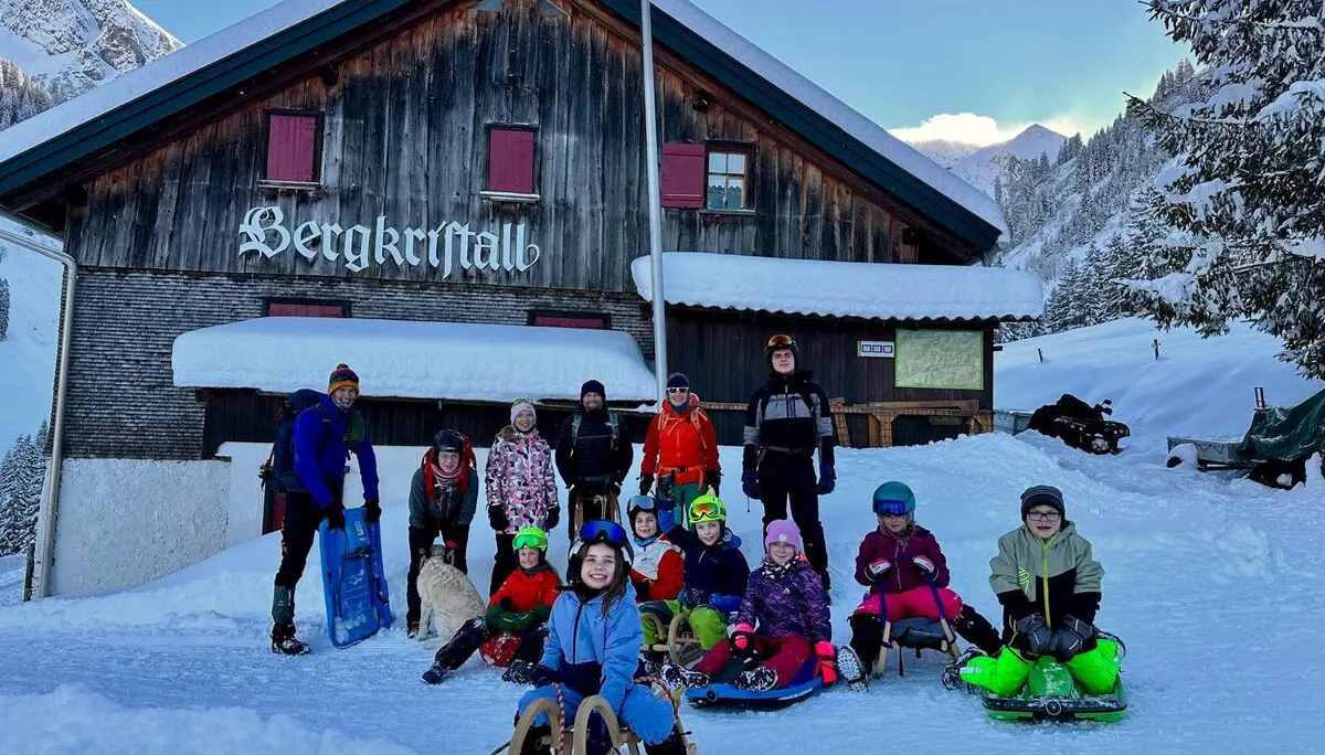 Gruppe von Kindern und Erwachsenen mit Schlitten vor einer Berghütte namens 'Bergkristall' in verschneiter Berglandschaft | © DAV-FN / A. Schababerle