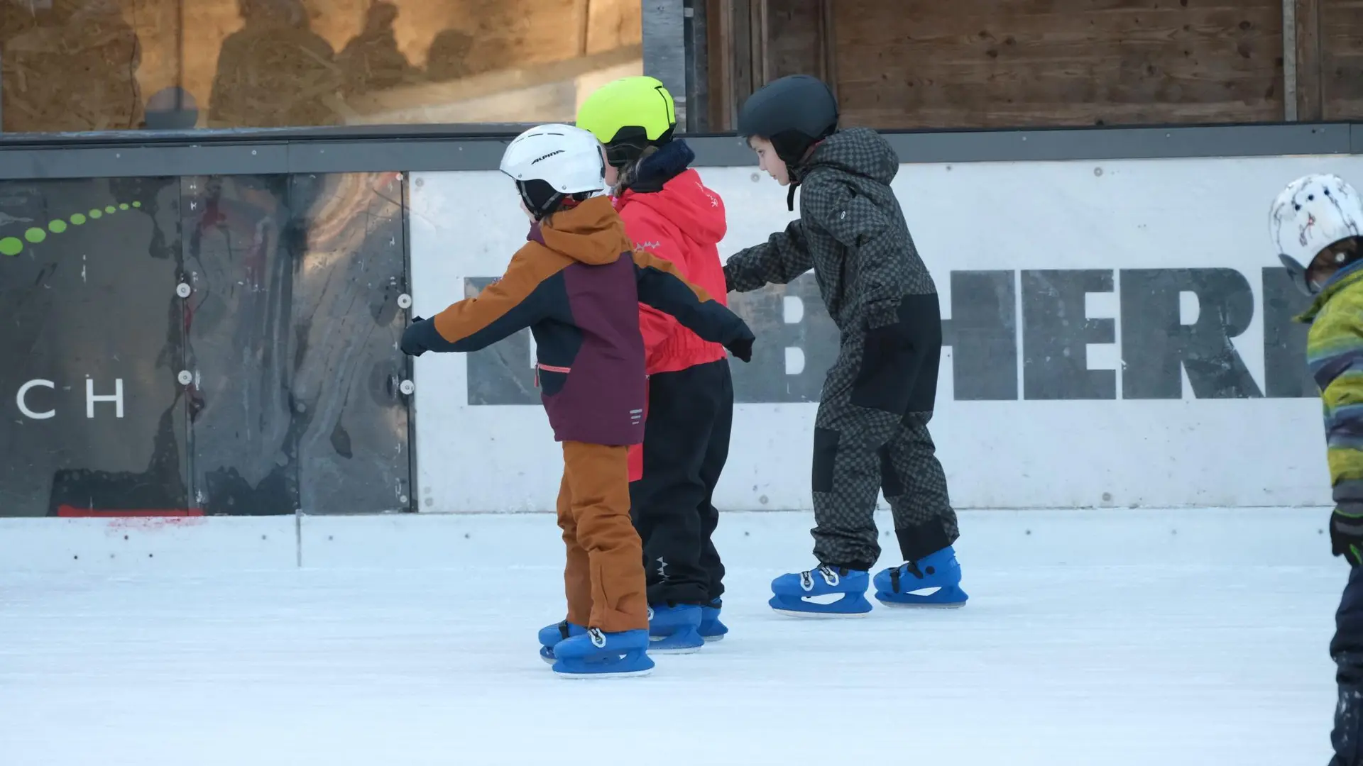 Drei Kinder mit Helmen und Schlittschuhen stehen nebeneinander auf einer Eisfläche und halten sich aneinander fest | © DAV-FN / A. Schababerle