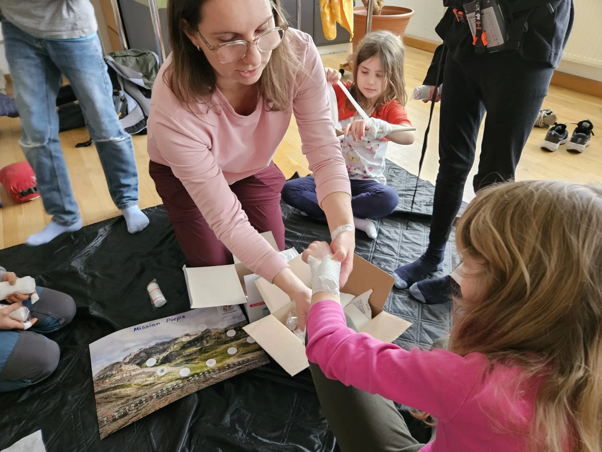 Mehrere Personen sitzen auf dem Boden, eine Person verbindet den Arm eines Kindes mit einer Bandage | © DAV-FN / A. Schababerle