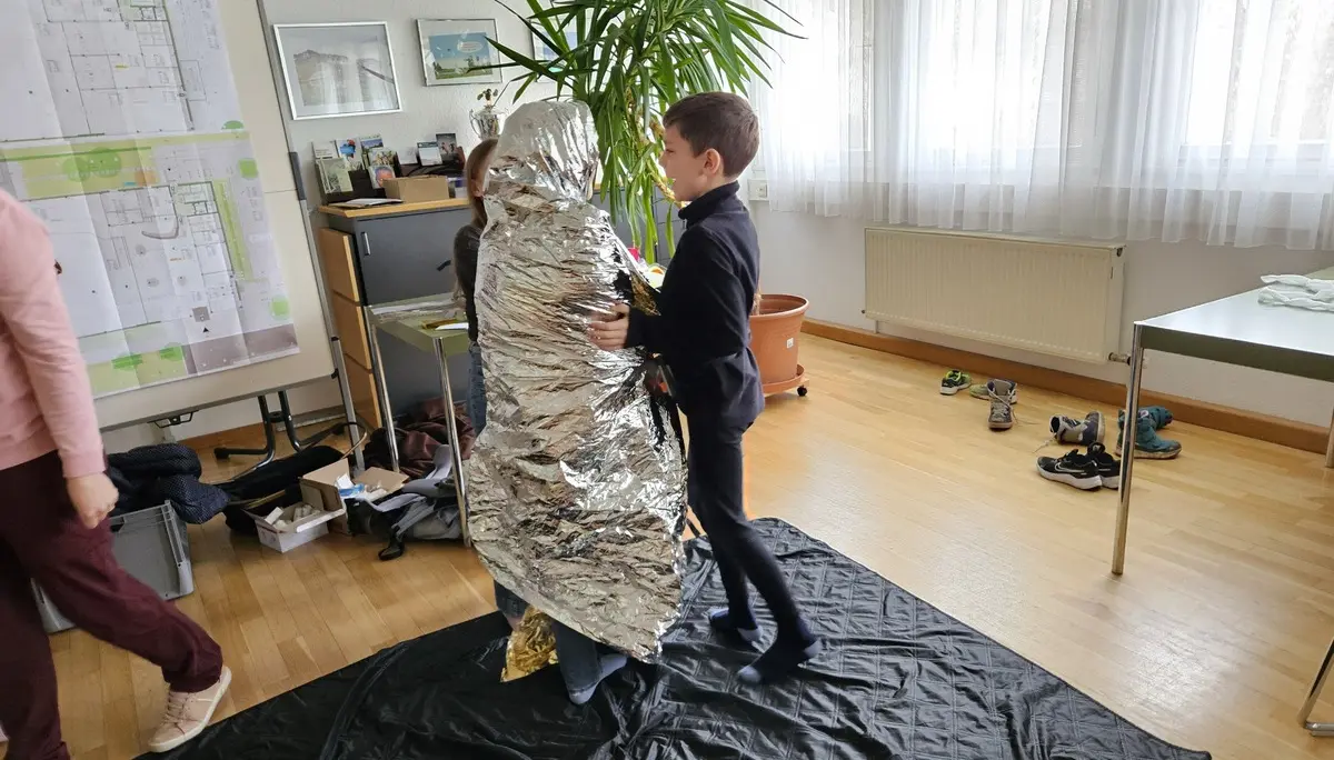 Kind steht vor einer Person, die in eine silberne Rettungsdecke eingewickelt ist, in einem hellen Raum mit Pflanzen und Möbeln | © DAV-FN / A. Schababerle