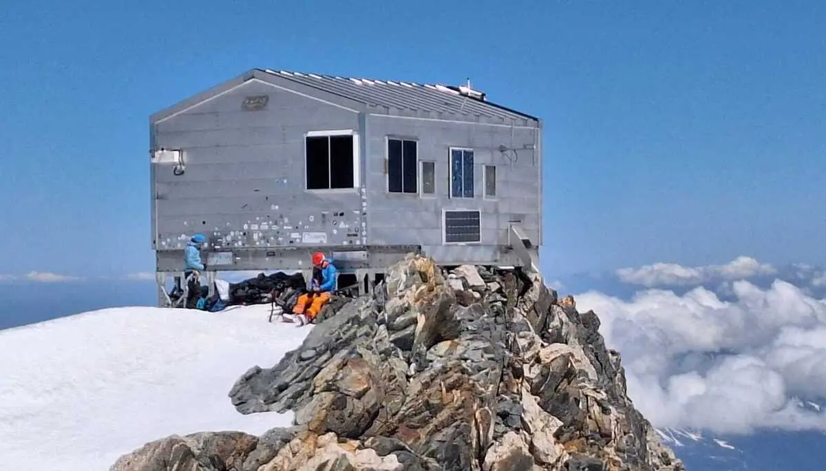 Holzhaus auf felsigem Gipfel über einer Wolkendecke mit zwei Personen in Bergsteigerkleidung davor | © DAV-FN / R. Gersten