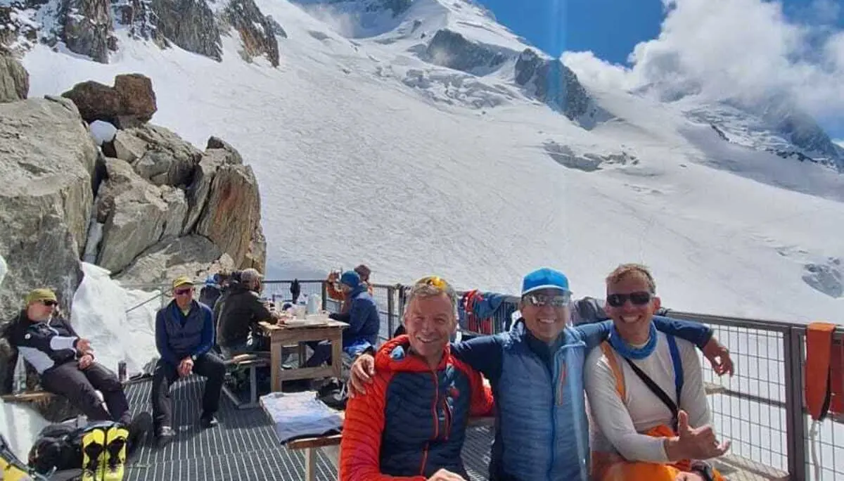 Drei Personen sitzen auf einer Metallgitterplattform vor schneebedeckten Bergen unter blauem Himmel mit Sonne | © DAV-FN / R. Gersten