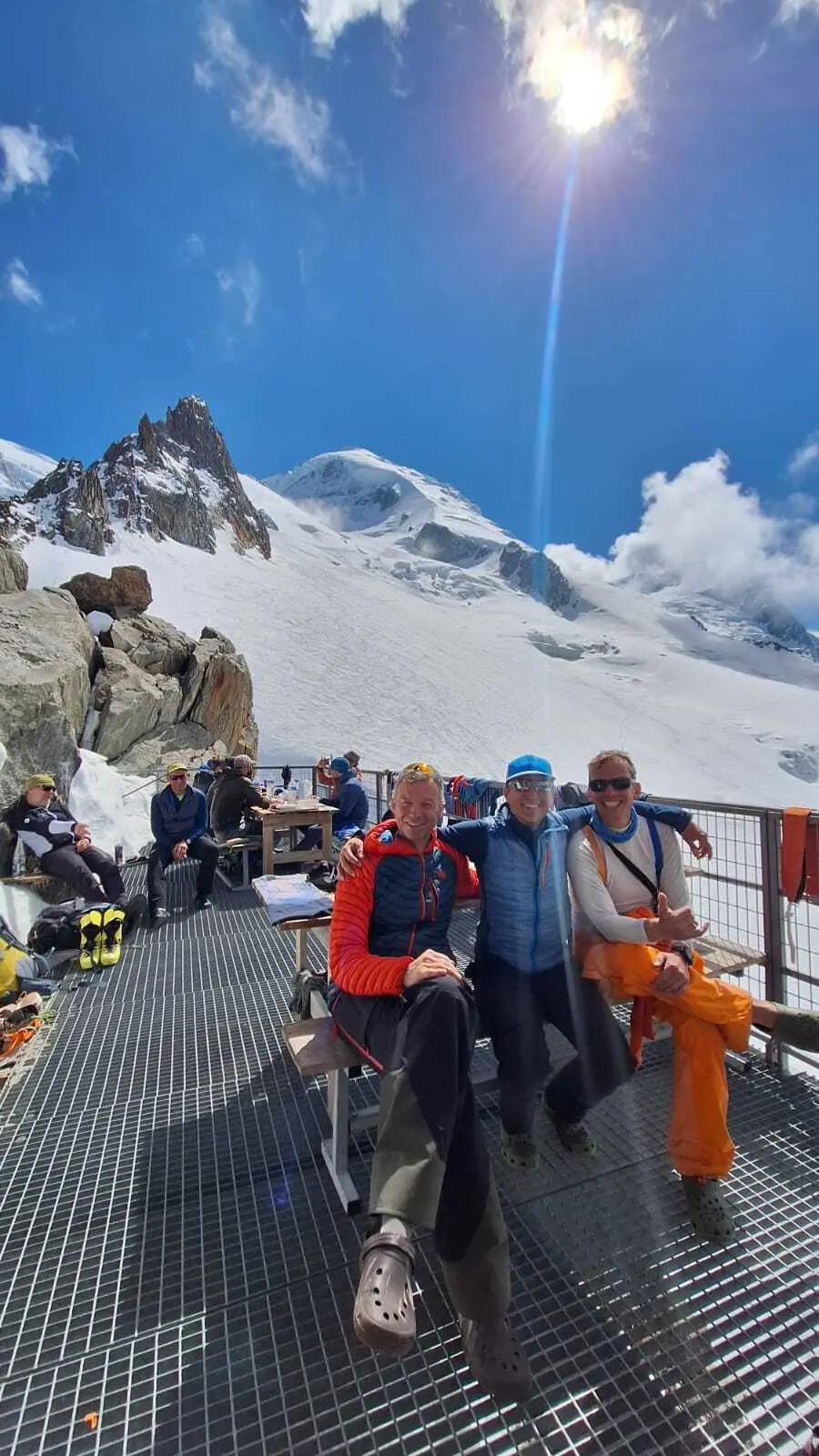 Drei Personen sitzen auf einer Metallgitterplattform vor schneebedeckten Bergen unter blauem Himmel mit Sonne | © DAV-FN / R. Gersten