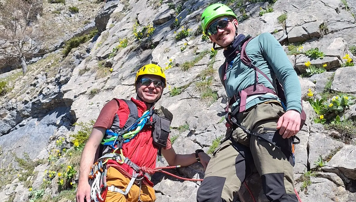 Zwei Männer mit Kletterausrüstung stehen an einem Felsen unter blauem Himmel | © DAV-FN / R. Huber