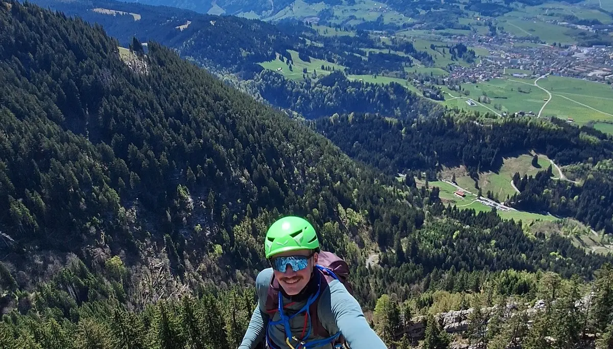 Kletterer mit grünem Helm und blauem Oberteil erklimmt felsigen Berggrat mit Blick auf bewaldete Hänge und Tal im Hintergrund | © DAV-FN / R. Huber