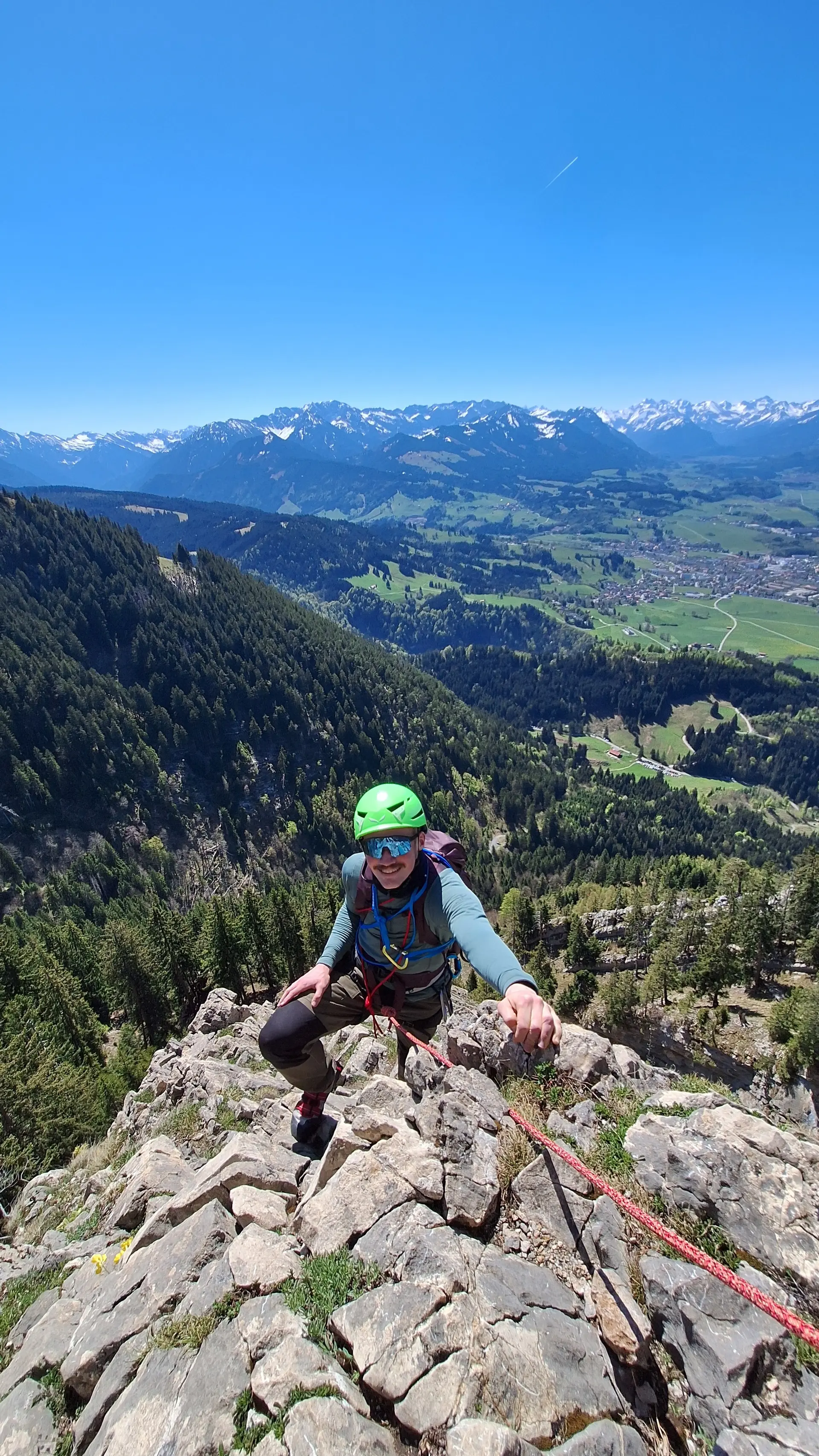 Kletterer mit grünem Helm und blauem Oberteil erklimmt felsigen Berggrat mit Blick auf bewaldete Hänge und Tal im Hintergrund | © DAV-FN / R. Huber