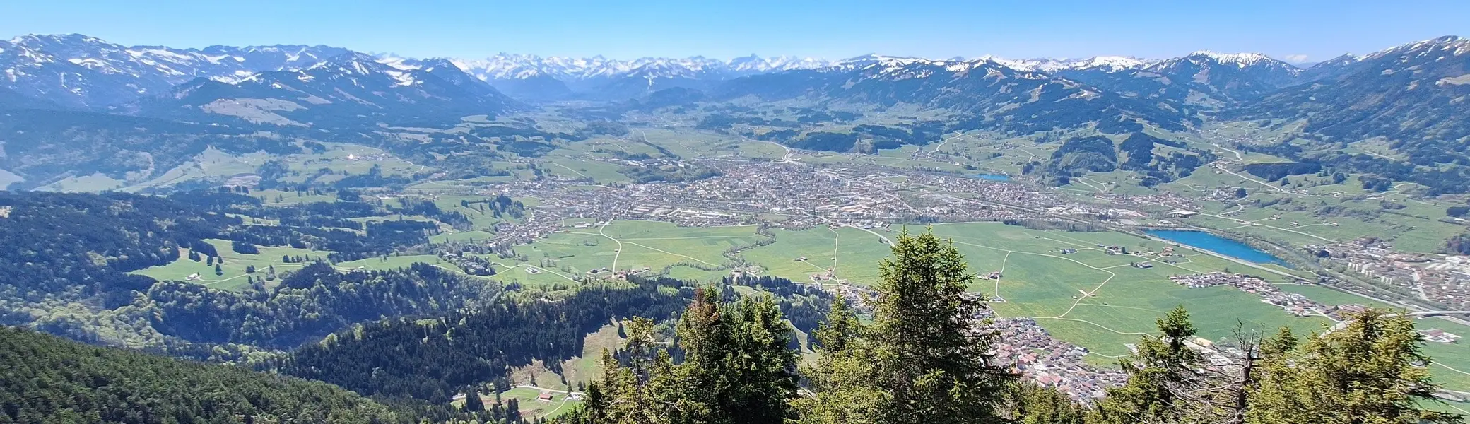 Blick von einem bewaldeten Berghang auf ein Tal mit einer Stadt und dahinter eine Bergkette mit schneebedeckten Gipfeln | © DAV-FN / R. Huber