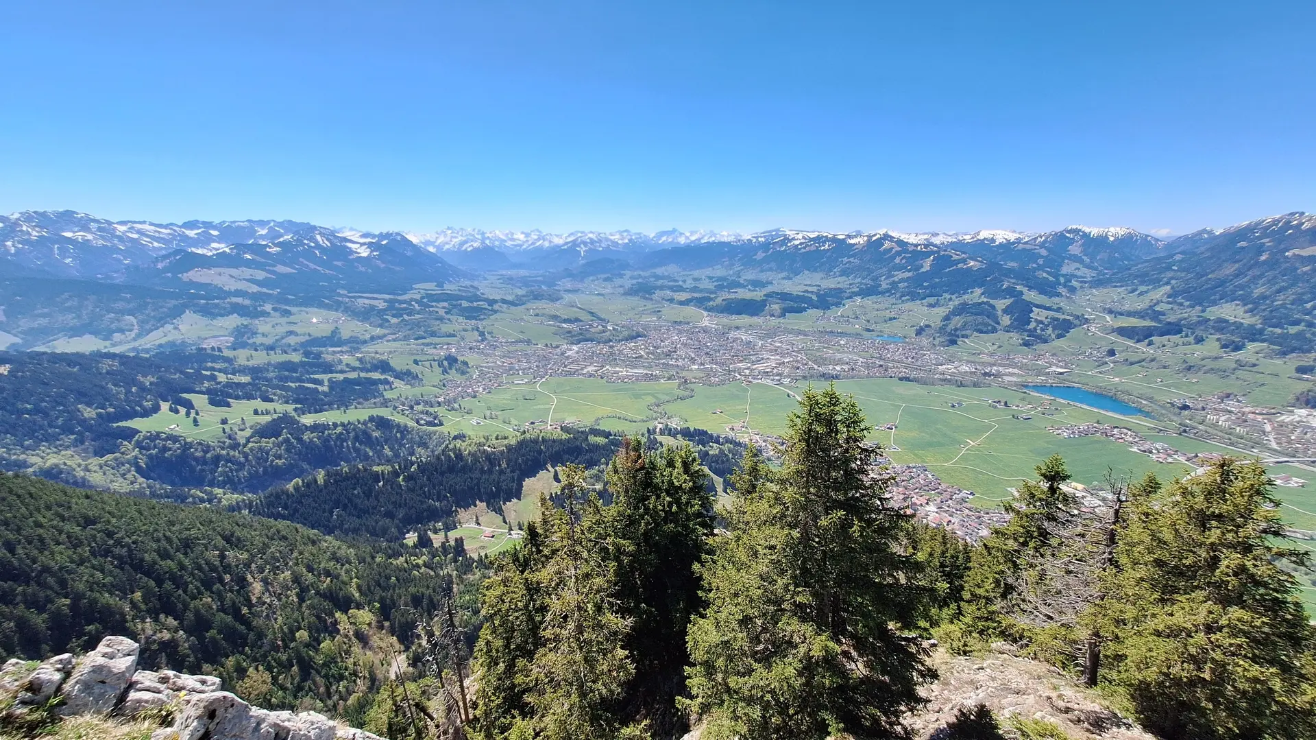 Blick von einem bewaldeten Berghang auf ein Tal mit einer Stadt und dahinter eine Bergkette mit schneebedeckten Gipfeln | © DAV-FN / R. Huber