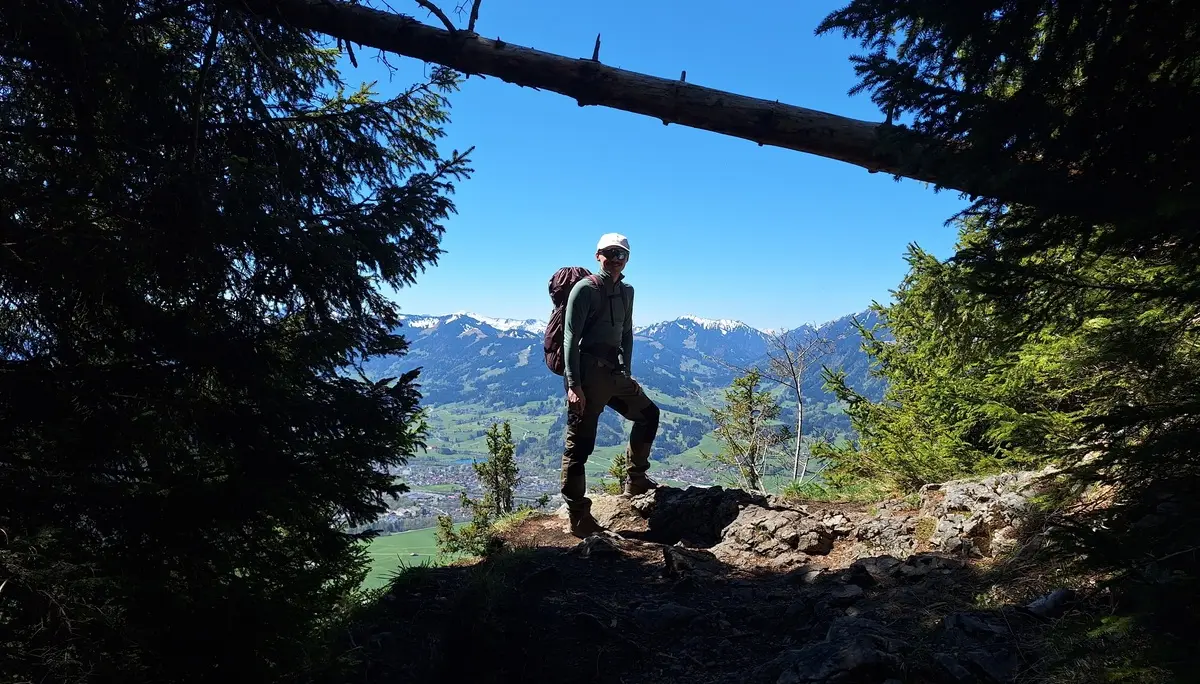 Wanderer mit Rucksack steht auf einem Felsen umgeben von Bäumen mit Bergpanorama im Hintergrund | © DAV-FN / R. Huber