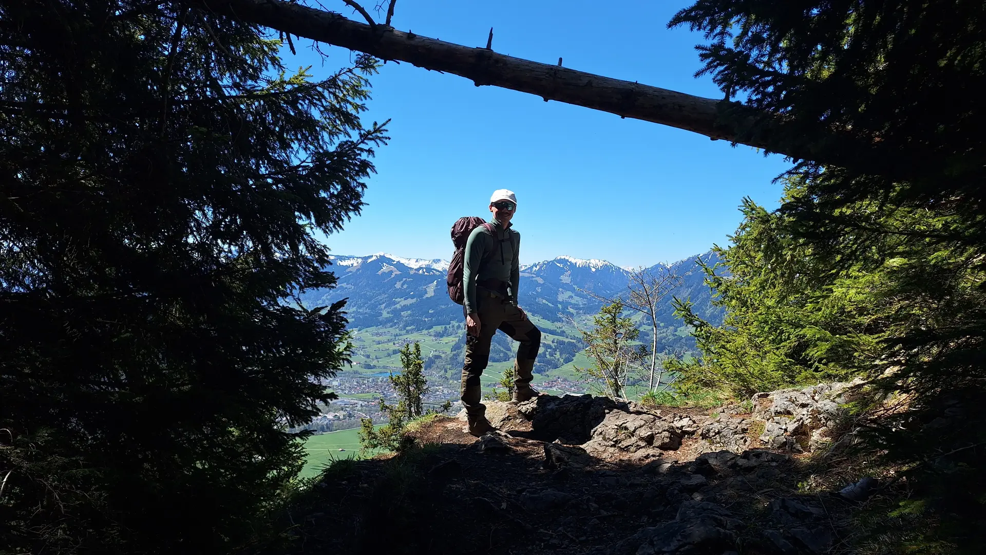 Wanderer mit Rucksack steht auf einem Felsen umgeben von Bäumen mit Bergpanorama im Hintergrund | © DAV-FN / R. Huber
