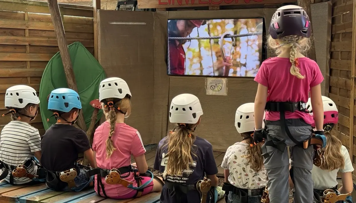 Sieben Kinder mit Kletterhelmen und Gurten sitzen auf einer Holzbank und schauen auf einen Bildschirm unter einem Schild mit der Aufschrift 'EINWEISUNG' | © DAV-FN / A. Schababerle