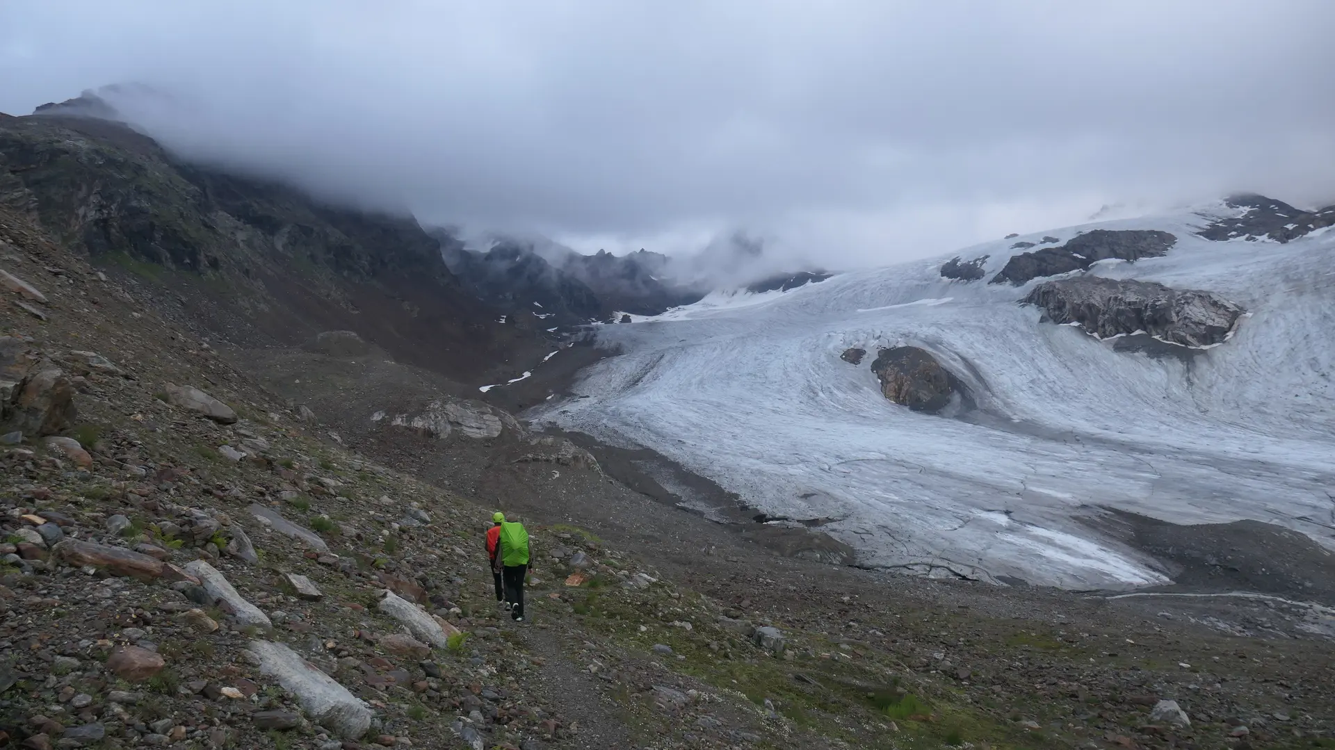Wanderer mit grünem Rucksack auf felsigem Pfad vor einem Gletscher unter bewölktem Himmel | © DAV - FN / S. Dischler