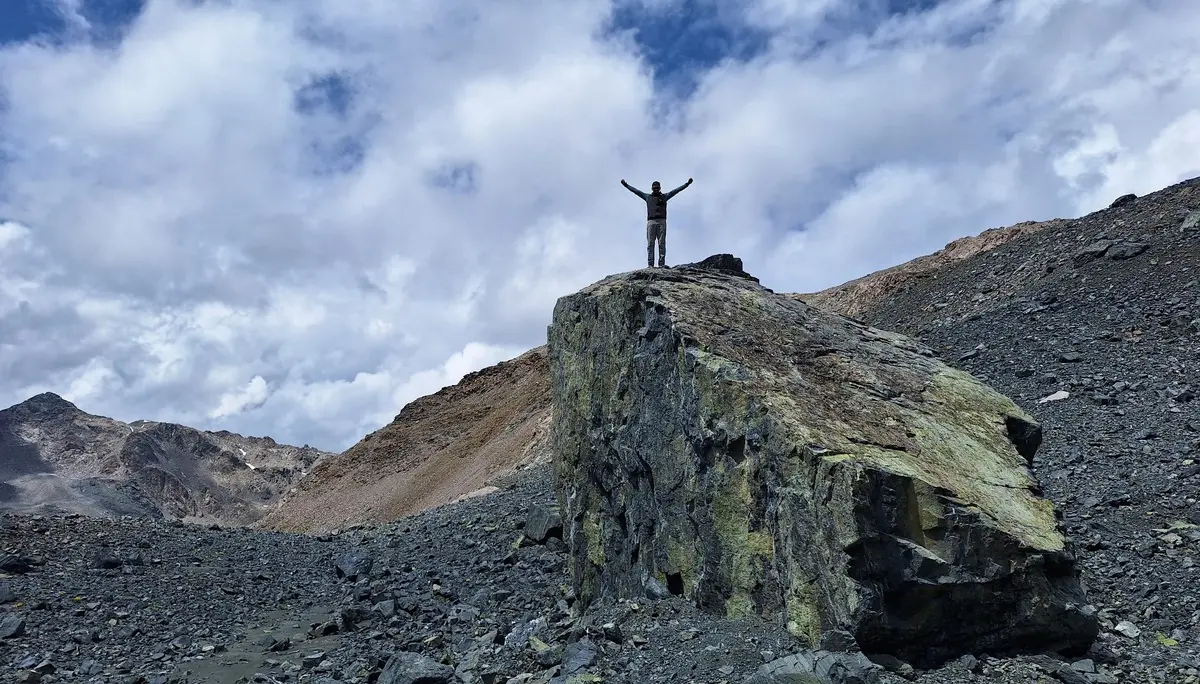 Person mit erhobenen Armen steht auf großem Felsen in bergiger Landschaft unter bewölktem Himmel | © Dav-Fn / R. Huber