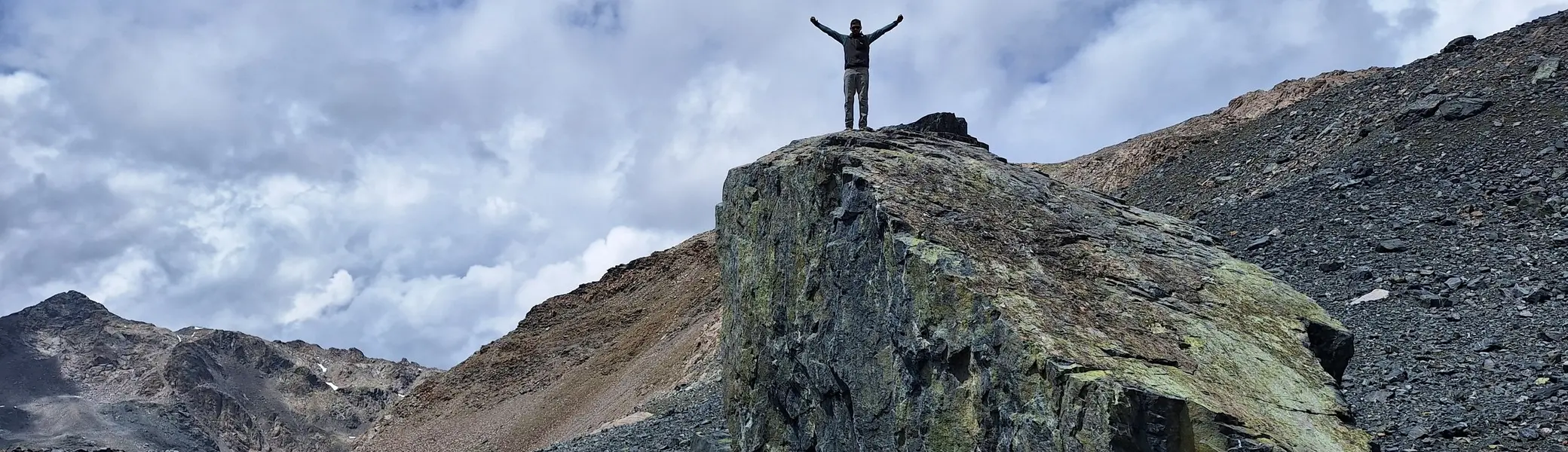 Person mit erhobenen Armen steht auf großem Felsen in bergiger Landschaft unter bewölktem Himmel | © Dav-Fn / R. Huber