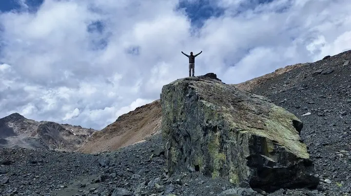 Person mit erhobenen Armen steht auf großem Felsen in bergiger Landschaft unter bewölktem Himmel | © Dav-Fn / R. Huber