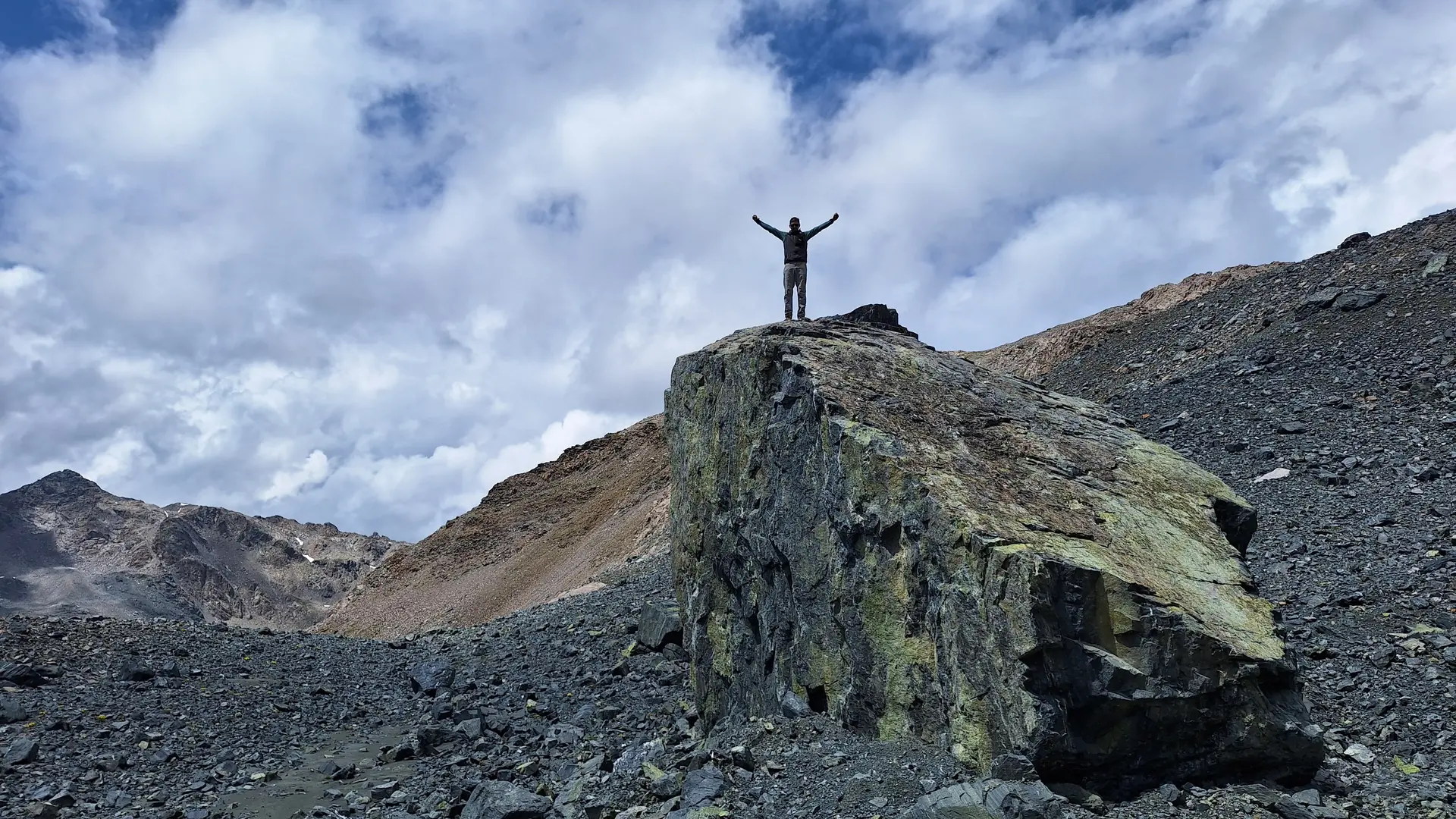 Person mit erhobenen Armen steht auf großem Felsen in bergiger Landschaft unter bewölktem Himmel | © Dav-Fn / R. Huber