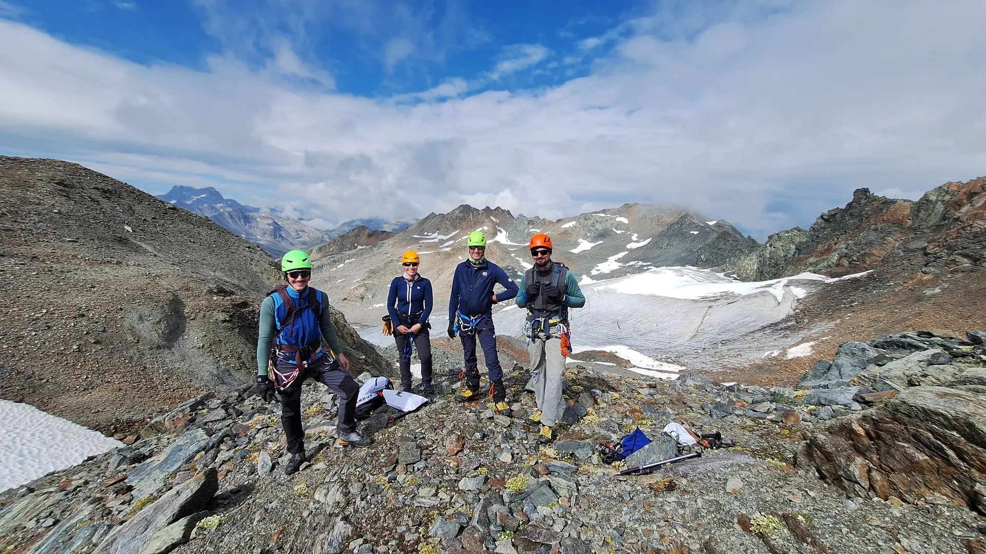 Vier Bergsteiger mit Helmen und Kletterausrüstung stehen auf felsigem Gipfel vor schneebedeckten Bergen unter blauem Himmel mit Wolken | © Dav-Fn / R. Huber
