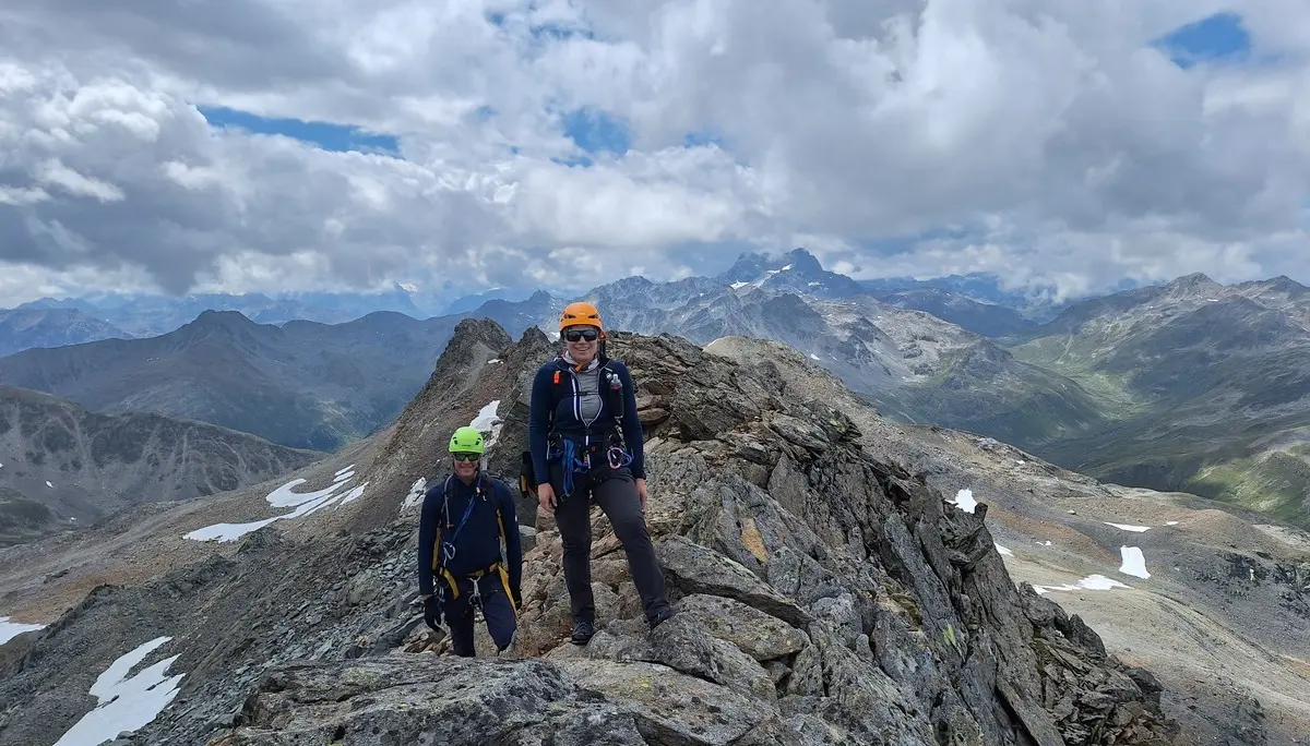 Zwei Bergsteiger mit Helmen und Rucksäcken auf felsigem Grat vor Bergpanorama unter bewölktem Himmel | © Dav-Fn / R. Huber