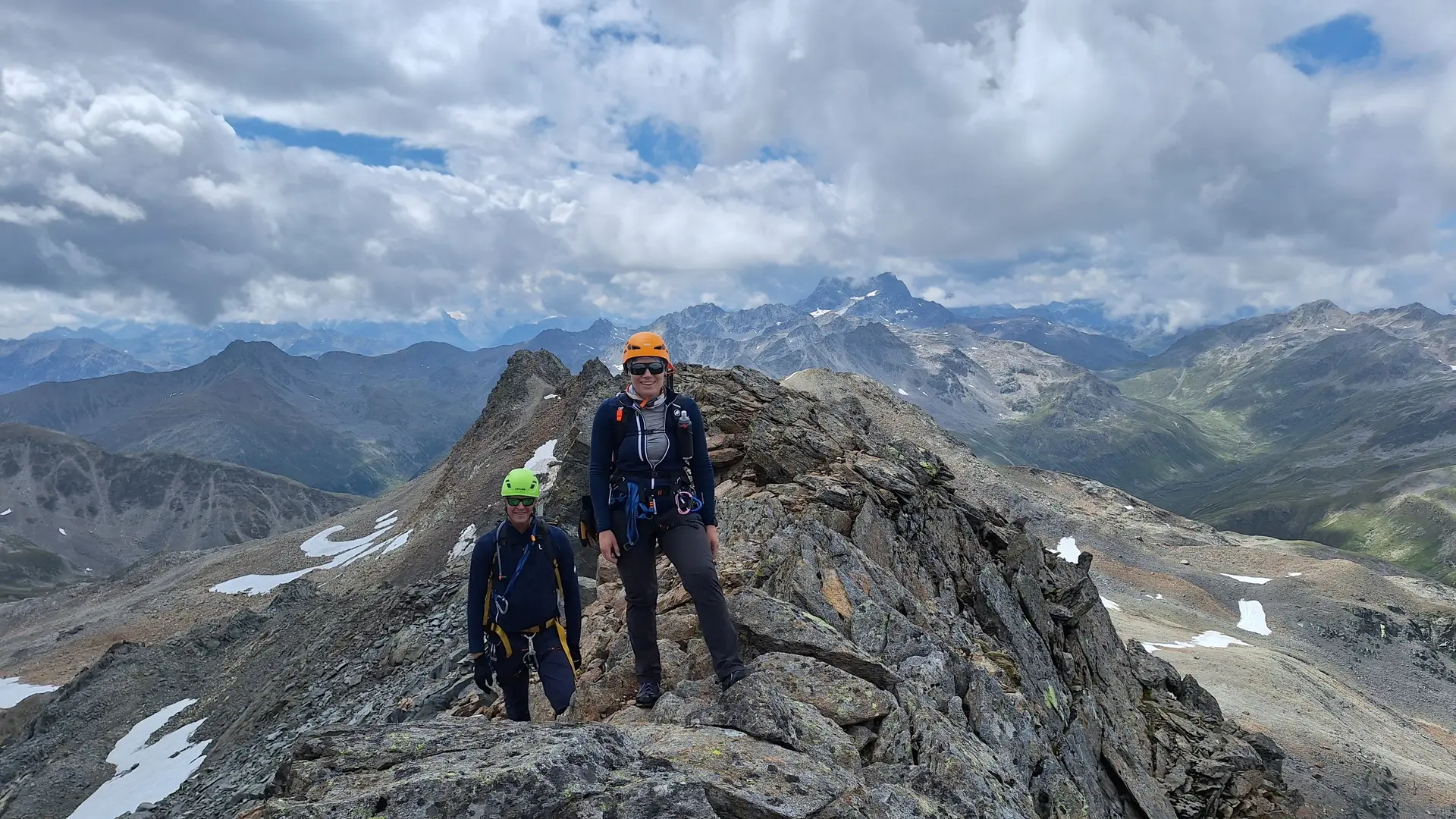 Zwei Bergsteiger mit Helmen und Rucksäcken auf felsigem Grat vor Bergpanorama unter bewölktem Himmel | © Dav-Fn / R. Huber