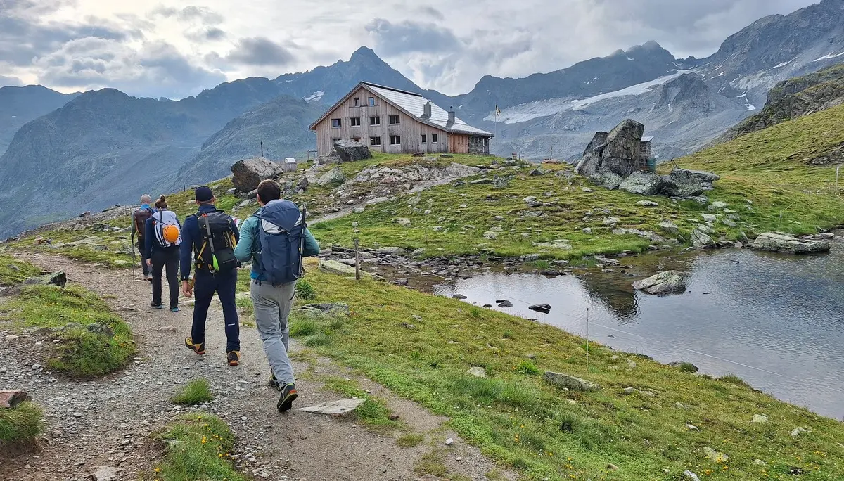 Gruppe von Wanderern auf einem Pfad in bergiger Landschaft mit See und Berghütte im Hintergrund | © Dav-Fn / R. Huber