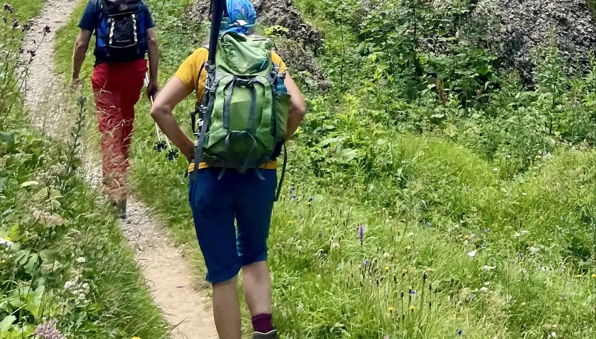 Zwei Wanderer mit Rucksäcken auf einem Pfad vor felsigem Hang mit Nadelbäumen unter bewölktem Himmel | © DAV-FN / K. Seuster