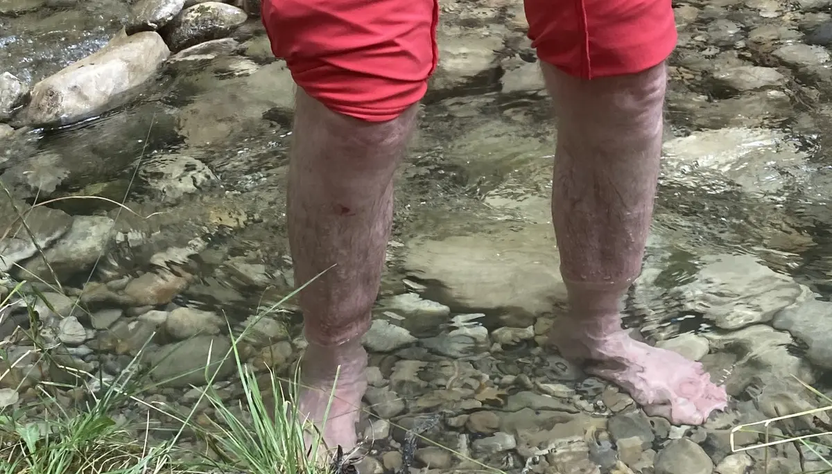 Beine einer Person in hochgekrempelter roter Hose stehen barfuß im flachen, klaren Bachwasser mit Steinen und Wurzeln | © DAV-FN / K. Seuster