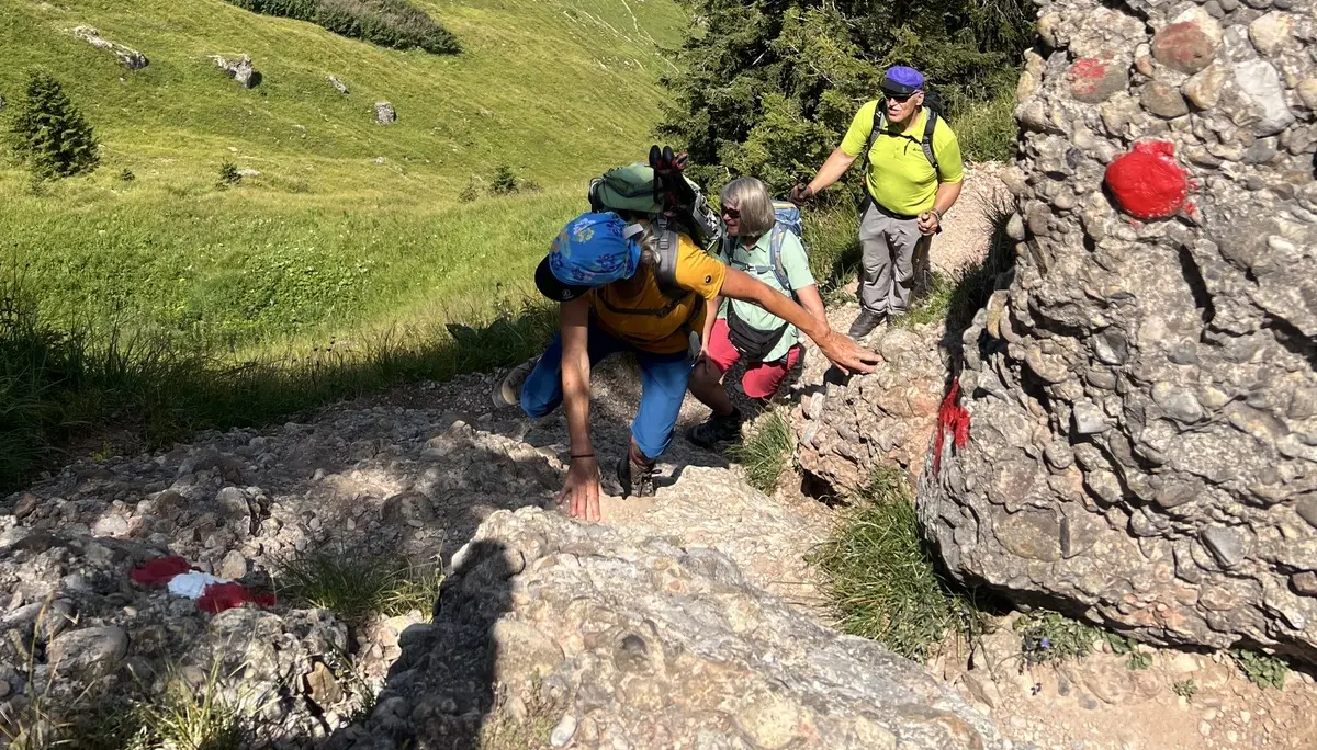 Drei Wanderer klettern an einem felsigen Hang mit roten Markierungen in einer grünen Berglandschaft | © DAV-FN / K. Seuster