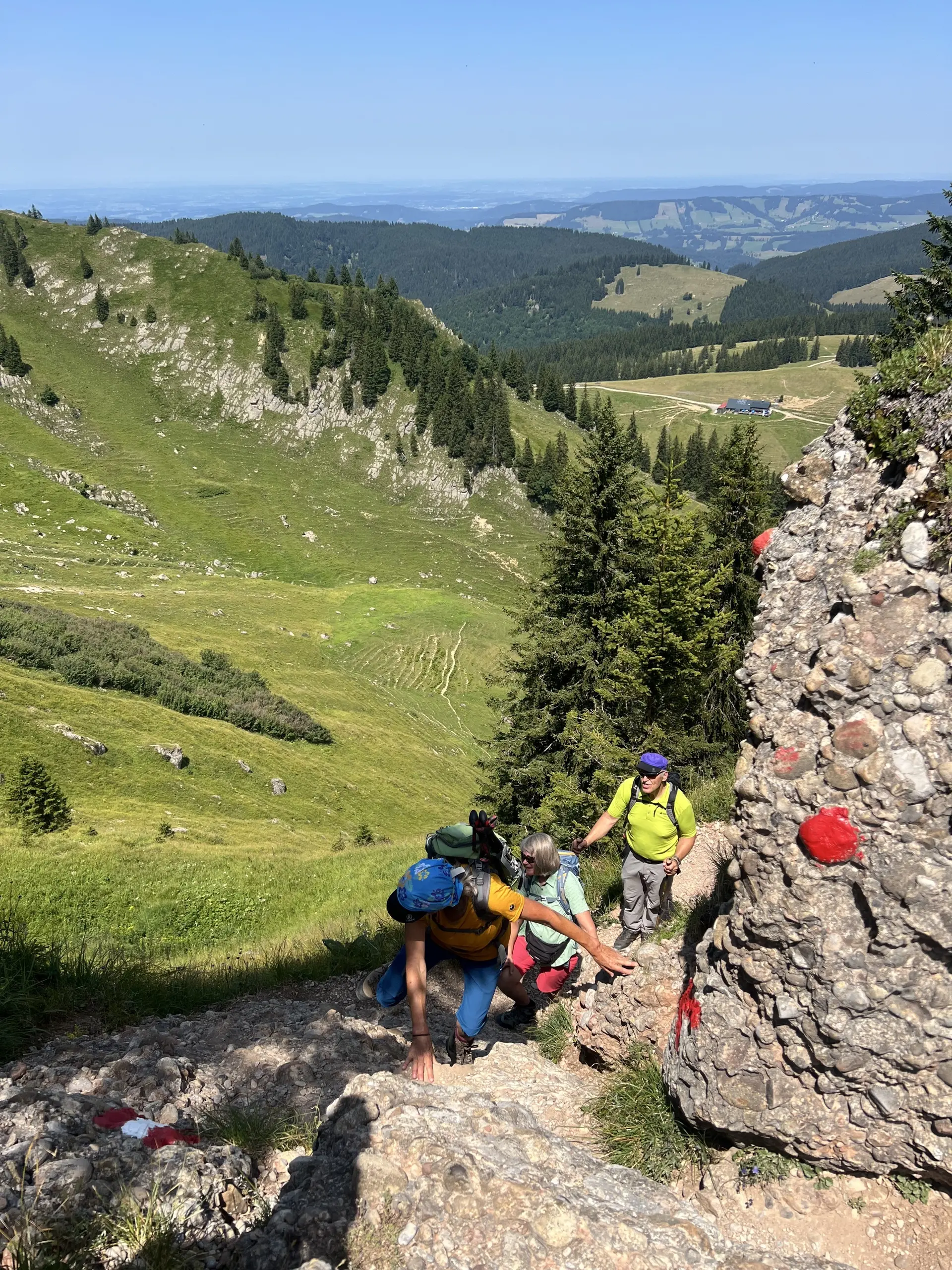 Drei Wanderer klettern an einem felsigen Hang mit roten Markierungen in einer grünen Berglandschaft | © DAV-FN / K. Seuster