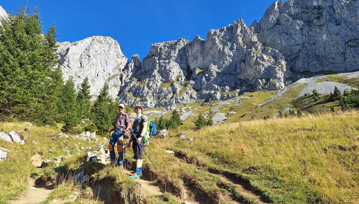 Zwei Wanderer mit Rucksäcken und Wanderstöcken auf einem Pfad vor felsigen Bergen und grüner Wiese | © DAV - FN / Yannic K., Robert H.