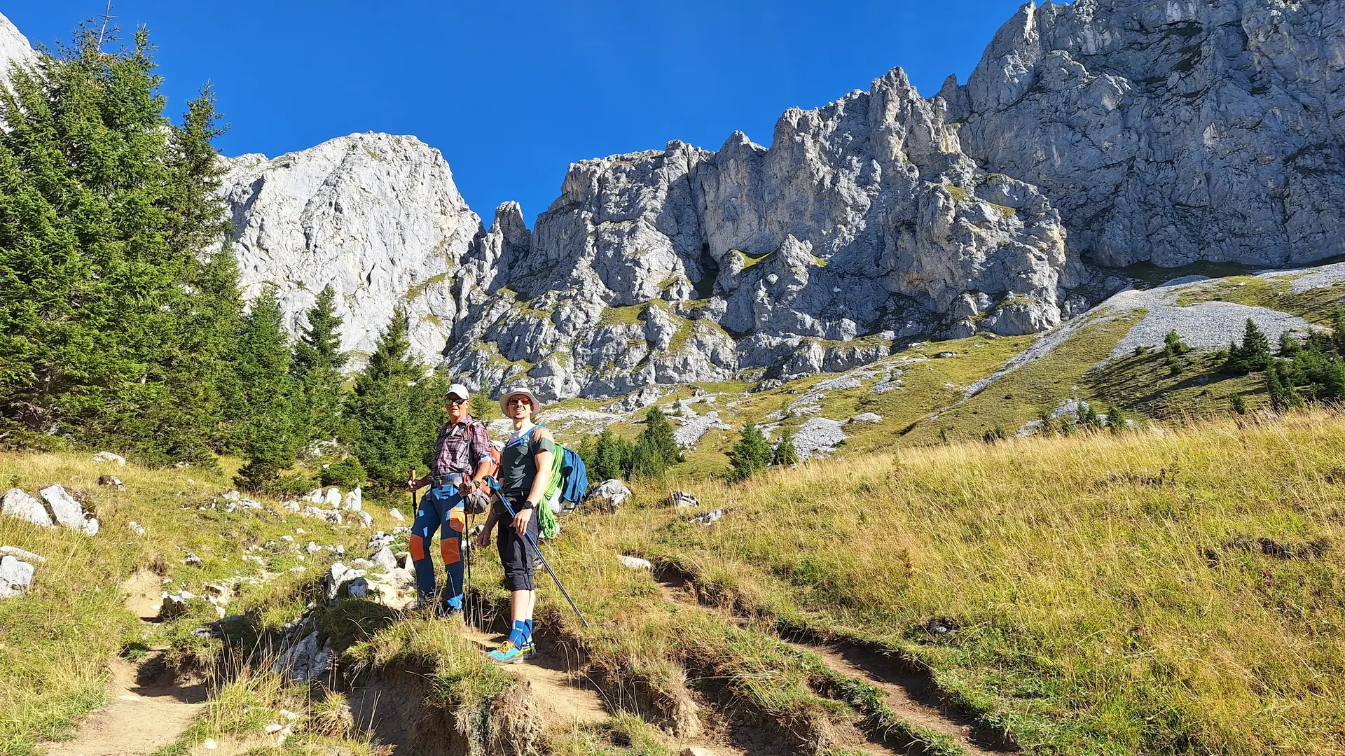 Zwei Wanderer mit Rucksäcken und Wanderstöcken auf einem Pfad vor felsigen Bergen und grüner Wiese | © DAV - FN / Yannic K., Robert H.
