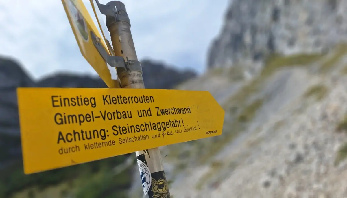 Gelber Wegweiser mit der Aufschrift 'Einstieg Kletterrouten Gimpel-Vorbau und Zwölferwand Achtung: Steinschlaggefahr' vor unscharfem Bergpanorama | © DAV - FN / Yannic K., Robert H.