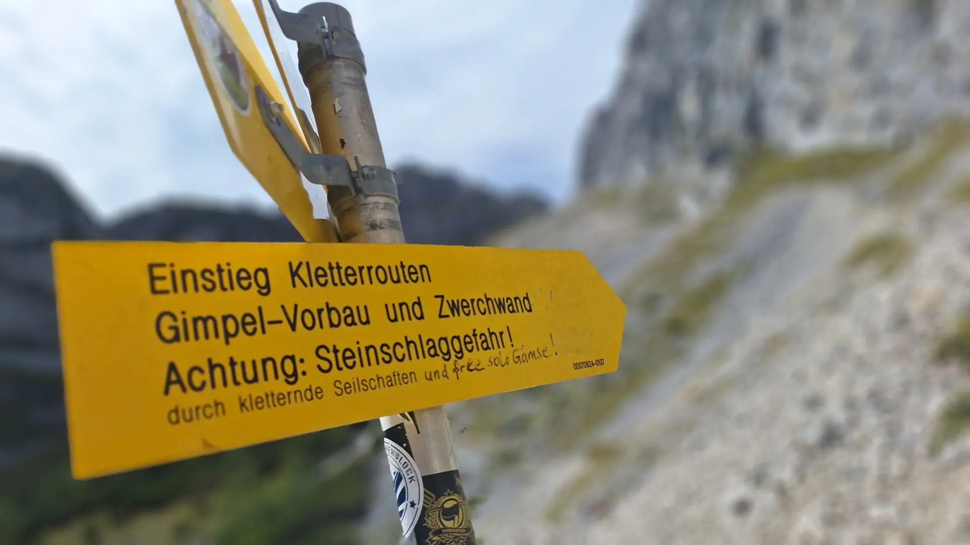 Gelber Wegweiser mit der Aufschrift 'Einstieg Kletterrouten Gimpel-Vorbau und Zwölferwand Achtung: Steinschlaggefahr' vor unscharfem Bergpanorama | © DAV - FN / Yannic K., Robert H.