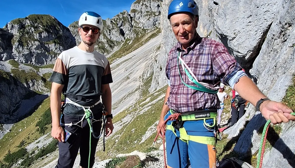 Zwei Männer mit Kletterausrüstung und Helmen stehen an einem Fels mit Berglandschaft im Hintergrund | © DAV - FN / Yannic K., Robert H.