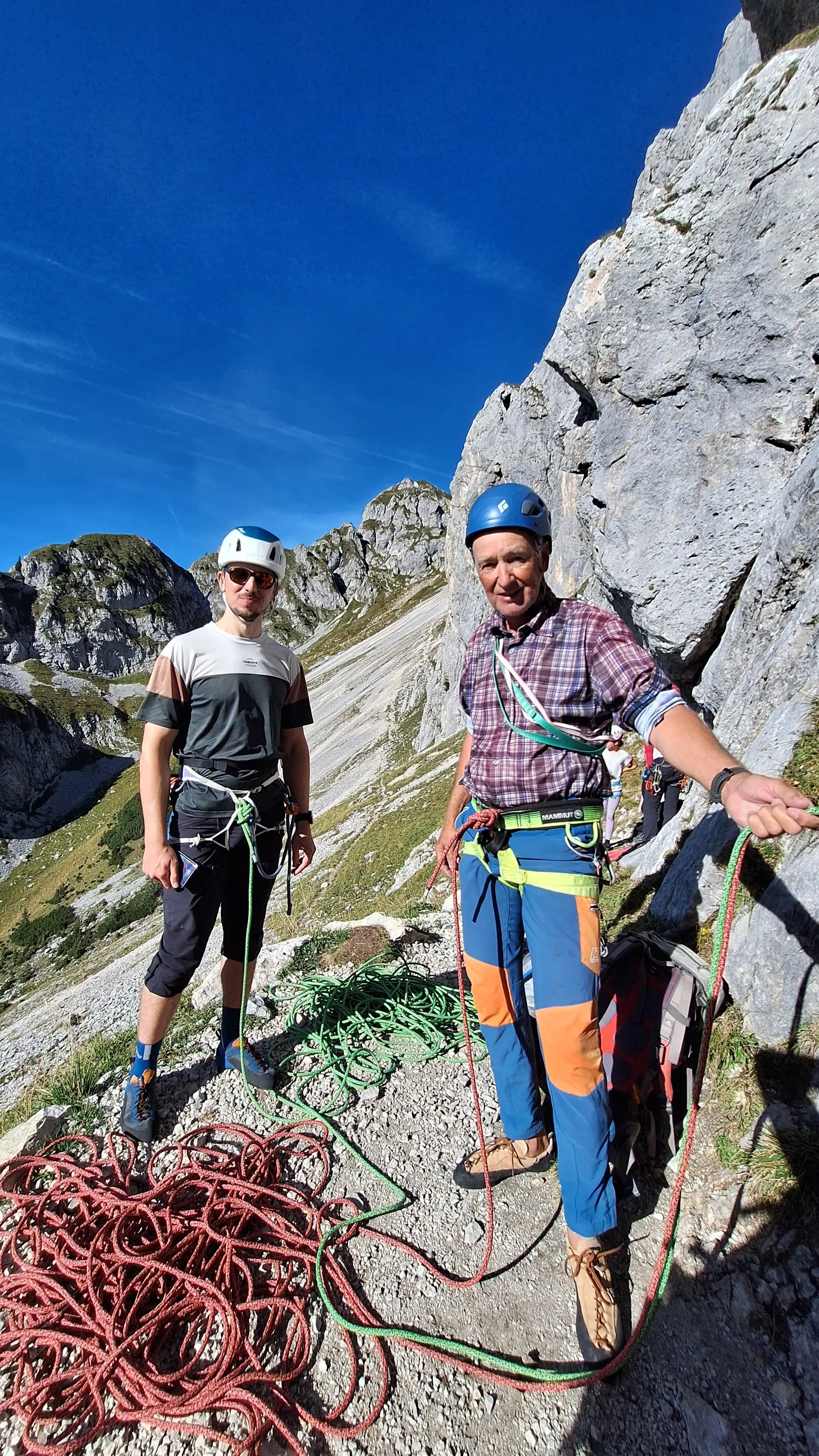 Zwei Männer mit Kletterausrüstung und Helmen stehen an einem Fels mit Berglandschaft im Hintergrund | © DAV - FN / Yannic K., Robert H.