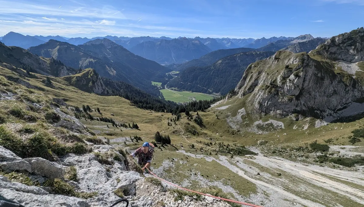 Bergsteiger mit Helm und Rucksack klettert an einem Seil an steilem Fels in einer alpinen Landschaft mit Tal und Bergen im Hintergrund | © DAV - FN / Yannic K., Robert H.