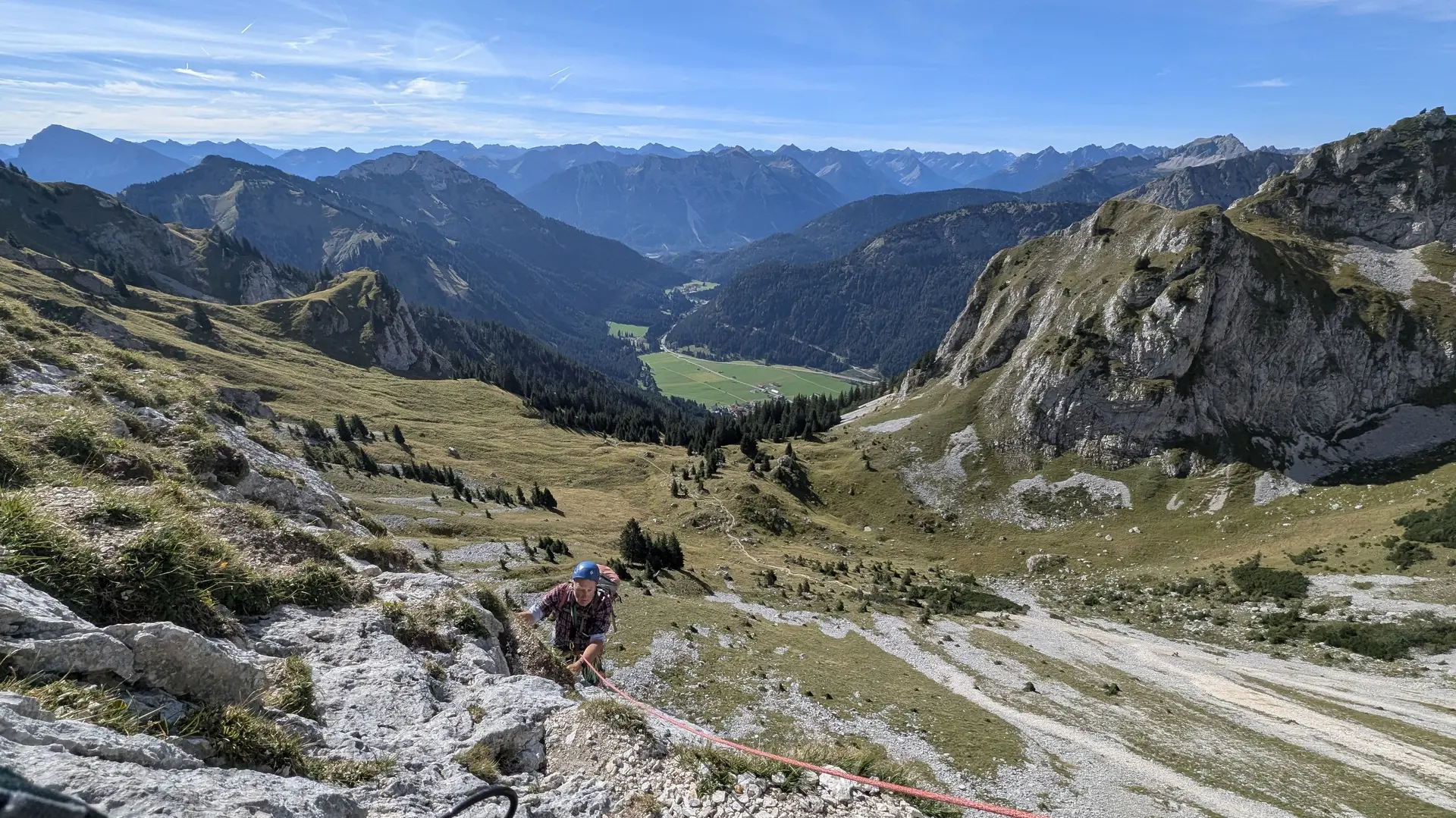 Bergsteiger mit Helm und Rucksack klettert an einem Seil an steilem Fels in einer alpinen Landschaft mit Tal und Bergen im Hintergrund | © DAV - FN / Yannic K., Robert H.
