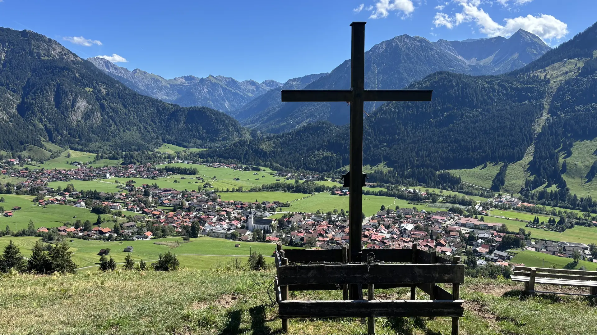 Holzkreuz und Holzbänke auf einer Wiese mit Blick auf ein Tal und Berge im Hintergrund | © DAV - FN / A. Schech