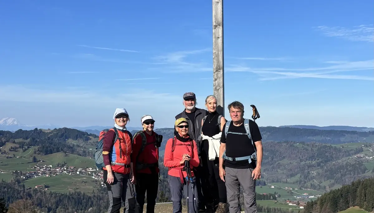 Sechs Wanderer stehen vor einem großen Holzkreuz auf dem Hündlekopf mit Blick auf eine ländliche Landschaft unter blauem Himmel | © DAV-FN / A. Schech