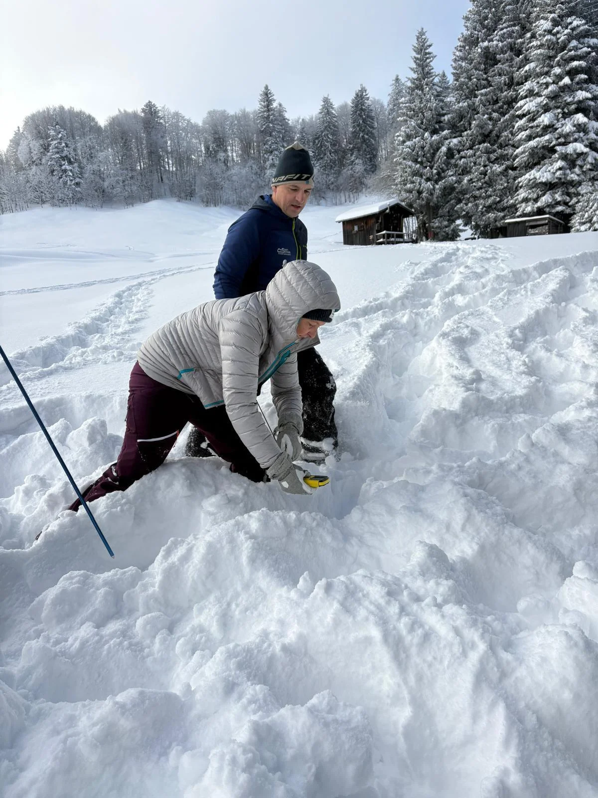 Zwei Personen knien im Schnee vor einem verschneiten Wald und einer Holzhütte, eine Person übt die Feinsuche mit dem LVS-Gerät | © DAV-FN / N. Gersten