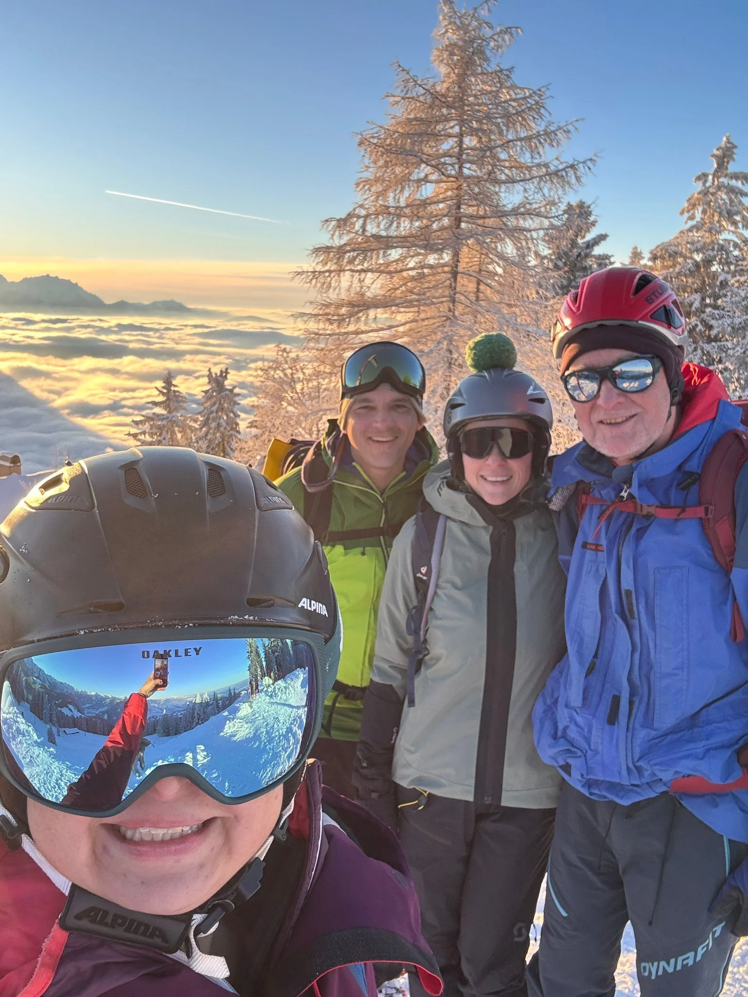 Gruppe von vier Personen mit Helmen und Outdoorbekleidung vor einer verschneiten Berglandschaft mit Baum und Wolkenmeer im Sonnenuntergang | © DAV-FN / Th. Gläser