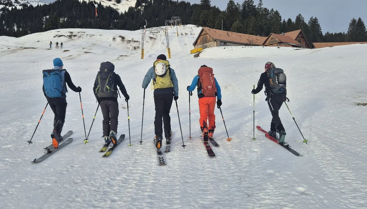 Fünf Skifahrer mit Rucksäcken steigen auf einer Schneepiste bergauf, im Hintergrund Hütten und Berge | © DAV-Fn / R. Lebedeff